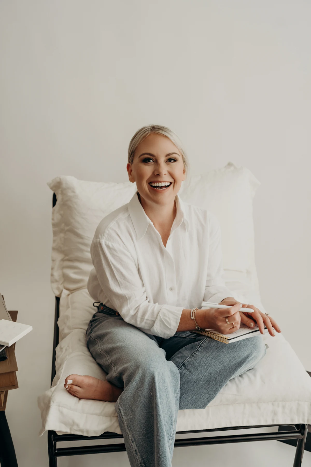 Smiling woman in a white shirt and jeans seated with a notebook and pen.