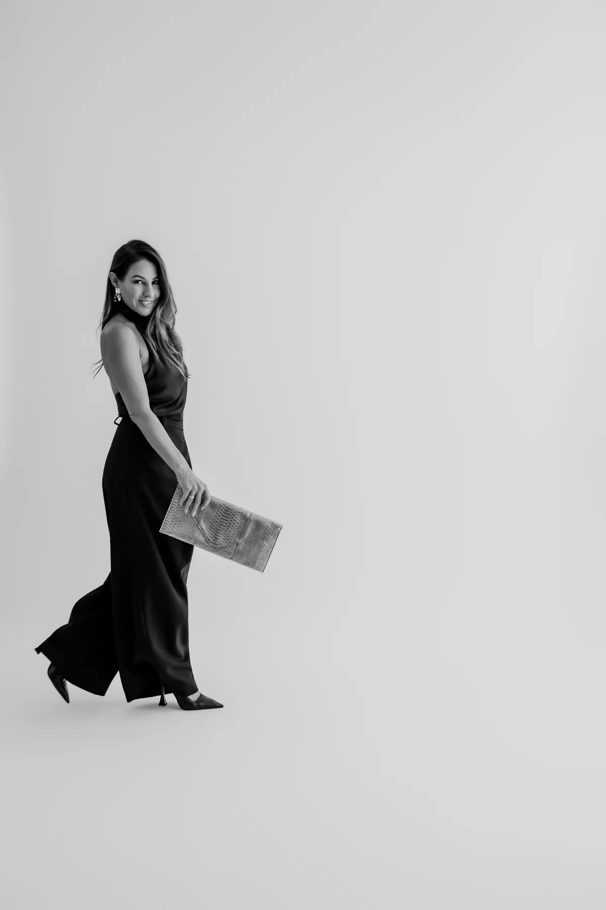 Black-and-white portrait of a woman walking in a black outfit while holding a clutch.