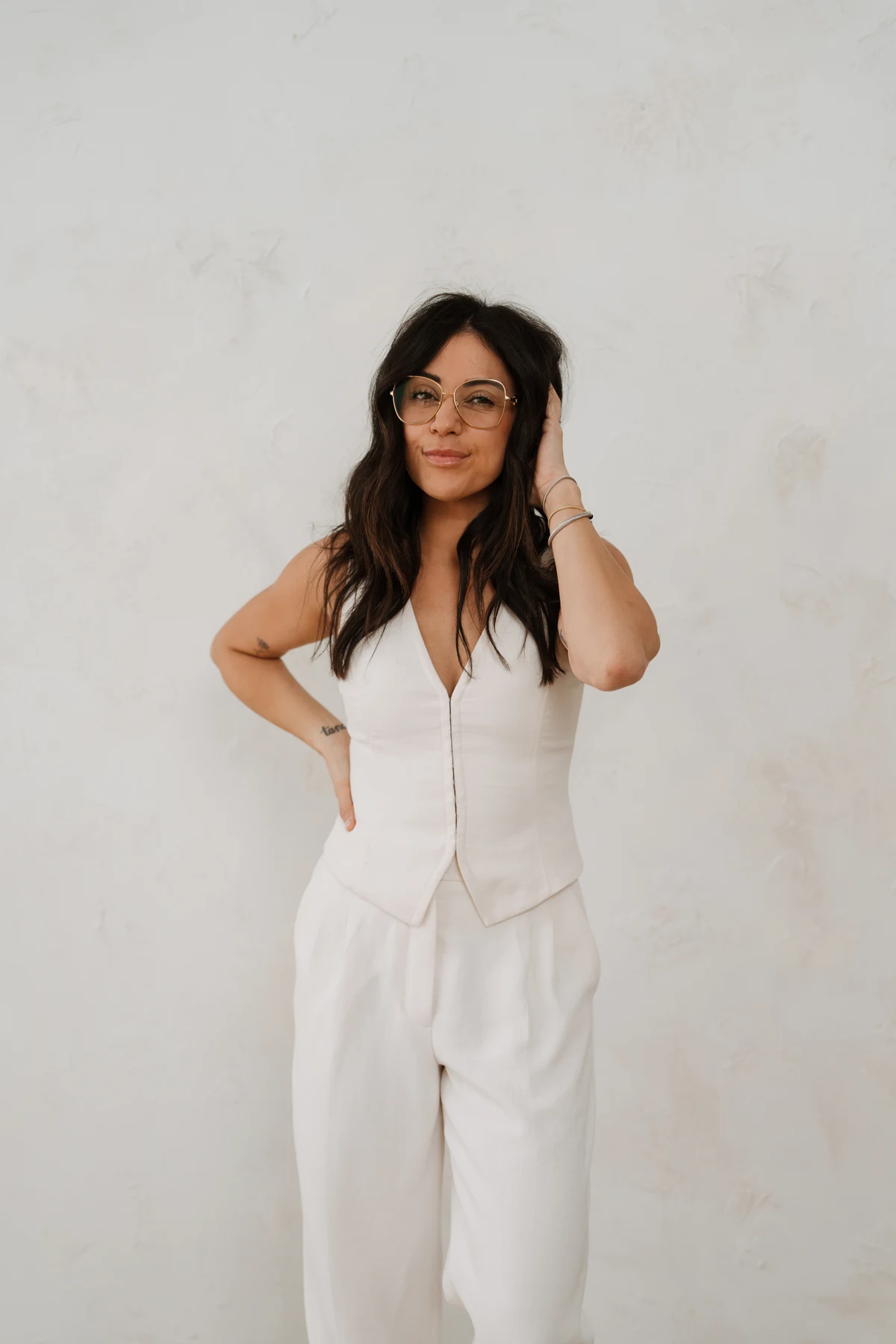 Jessica wearing glasses and a white vest and trousers against a textured studio wall.