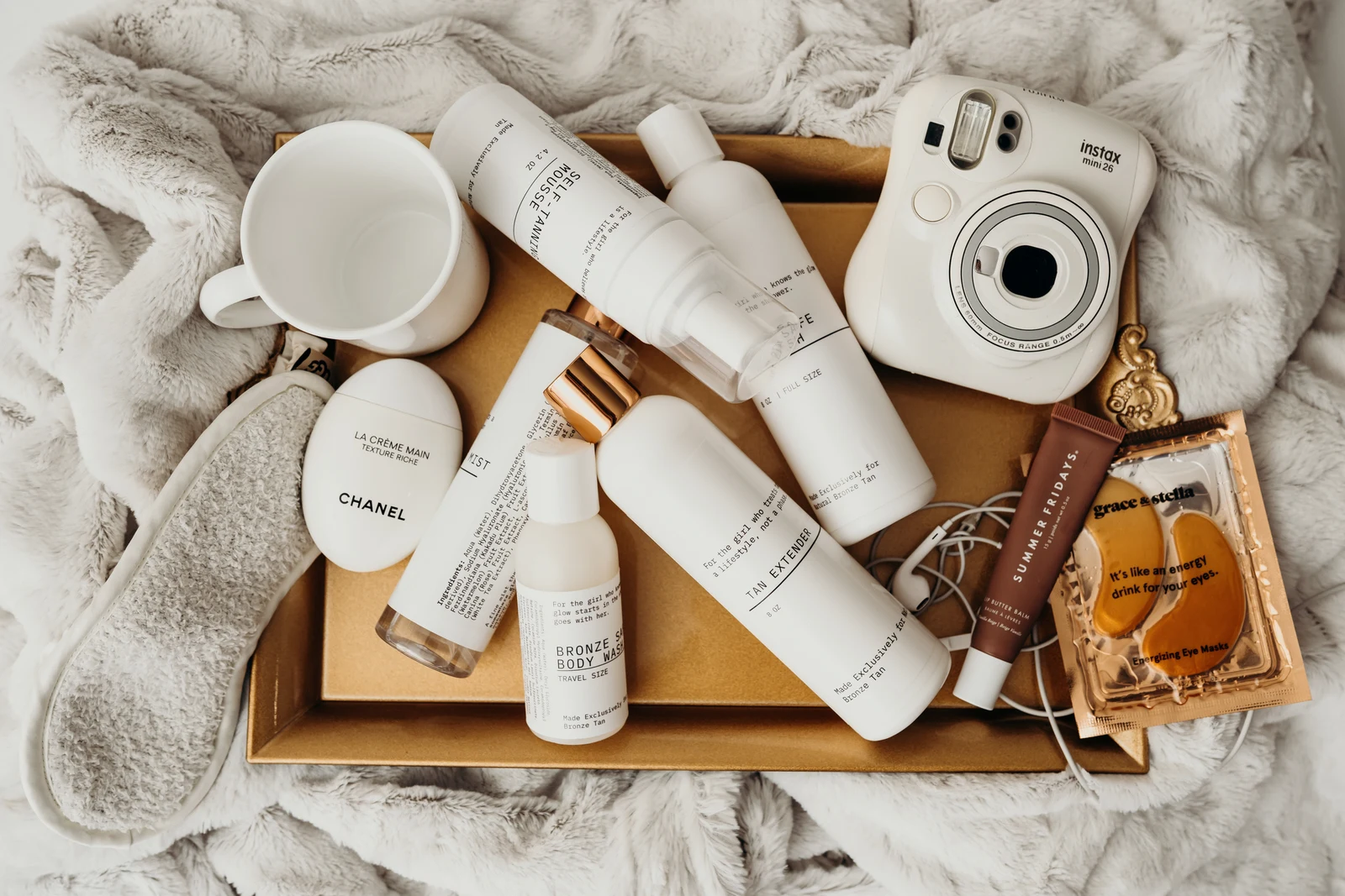 Beauty products, a white instant camera, and a cup arranged on a gold tray over a soft blanket.